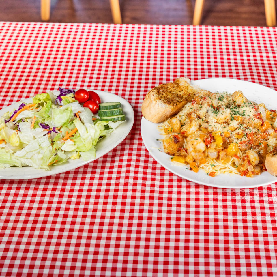 Plates of mixed salad and seafood pasta with toasted bread on a red-and-white checkered restaurant table.