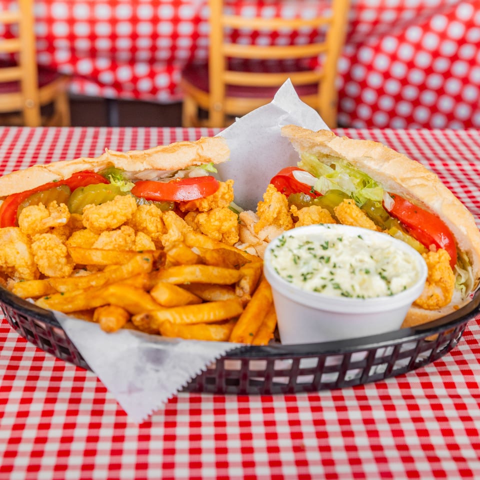 Basket of fried seafood sandwiches with lettuce, tomatoes, pickles, fries, and dipping sauce on a red-and-white checkered tablecloth.