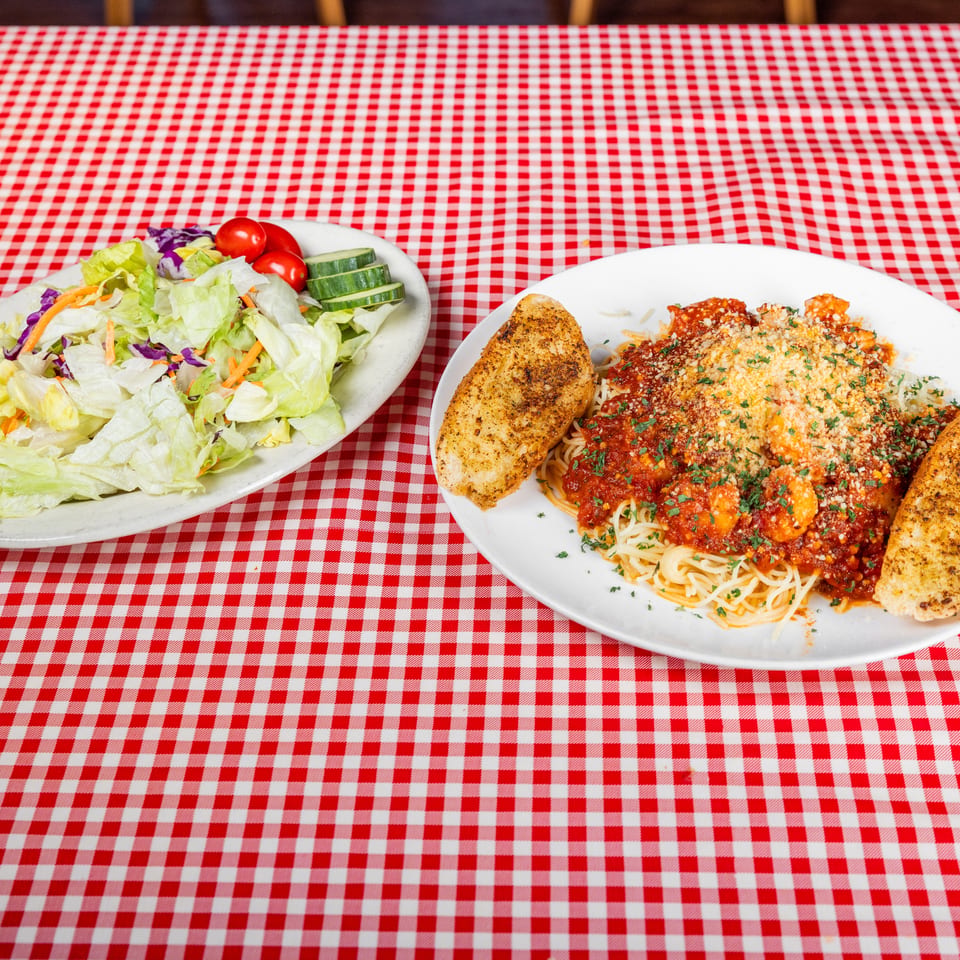 Spaghetti with marinara, shrimp, grilled chicken, and garlic bread beside a garden salad on a red-and-white checkered tablecloth.