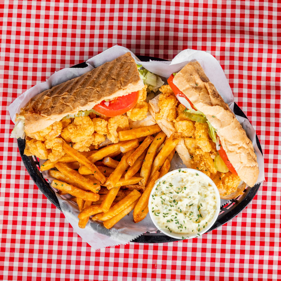 Basket of fried shrimp sandwiches with lettuce and tomatoes, French fries, and dipping sauce on a red-and-white checkered tablecloth.