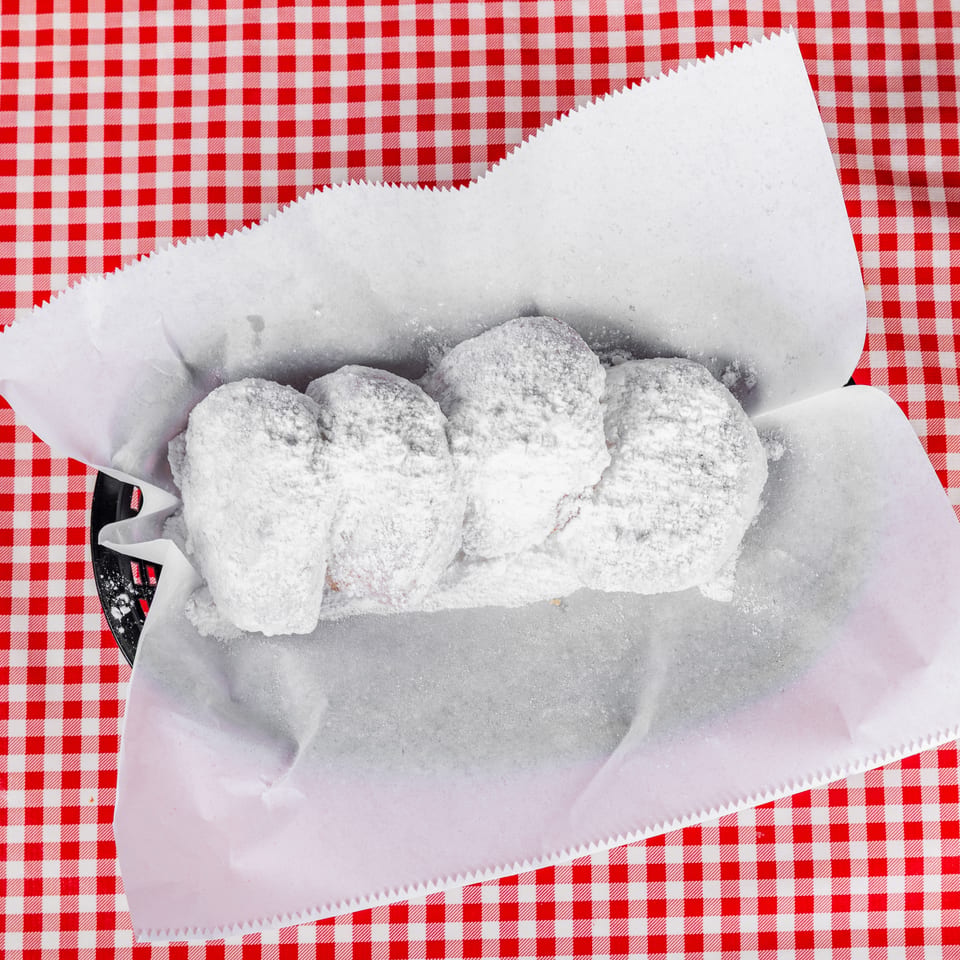 Powdered sugar-coated pastries served in a paper-lined basket on a red-and-white checkered tablecloth.