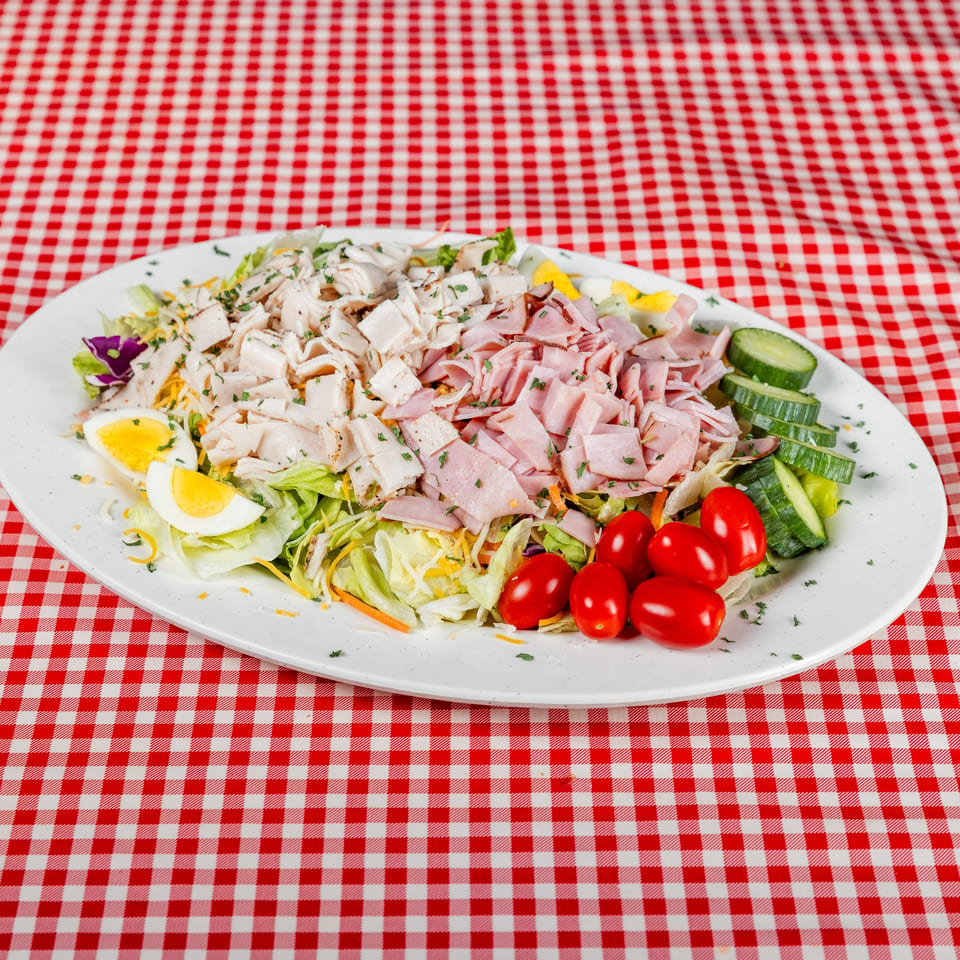 Chef salad with grilled chicken, ham, lettuce, eggs, cucumbers, tomatoes, and cheese on a red-checkered tablecloth.
