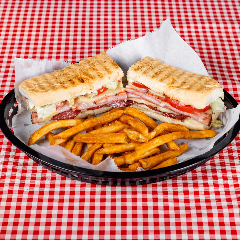 Two grilled deli sandwiches with ham, bacon, cheese, lettuce, and tomato served with fries on a black tray.