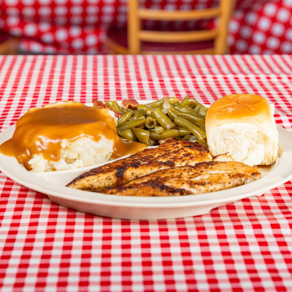 Plate of grilled chicken with mashed potatoes and gravy, green beans, and a dinner roll on a red-and-white checkered tablecloth.