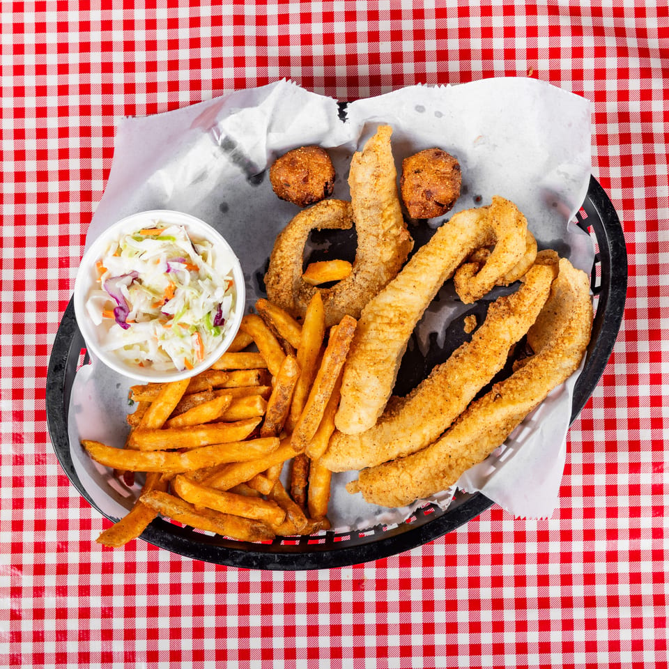 Basket of fried fish, shrimp, hush puppies, and fries with coleslaw on a red-and-white checkered tablecloth.