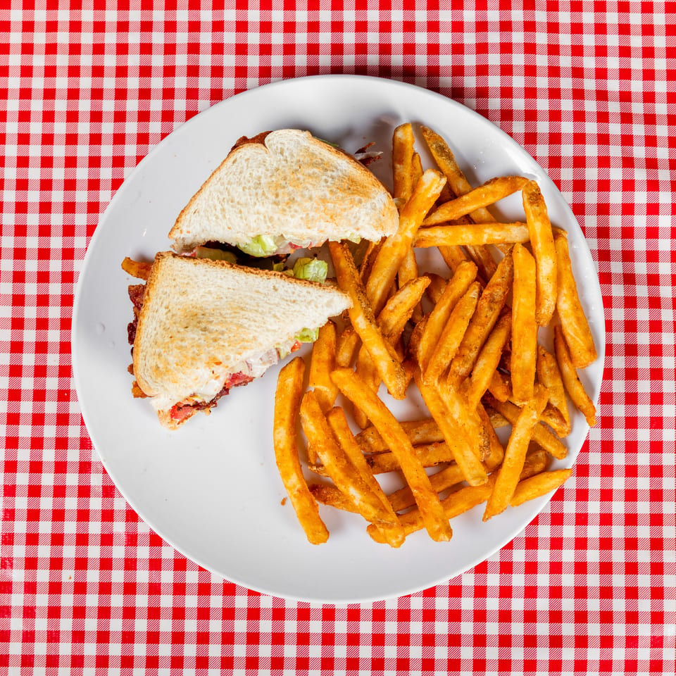 Toasted bacon, lettuce, and tomato sandwiches served with golden fries on a white plate over a red-and-white checkered tablecloth.