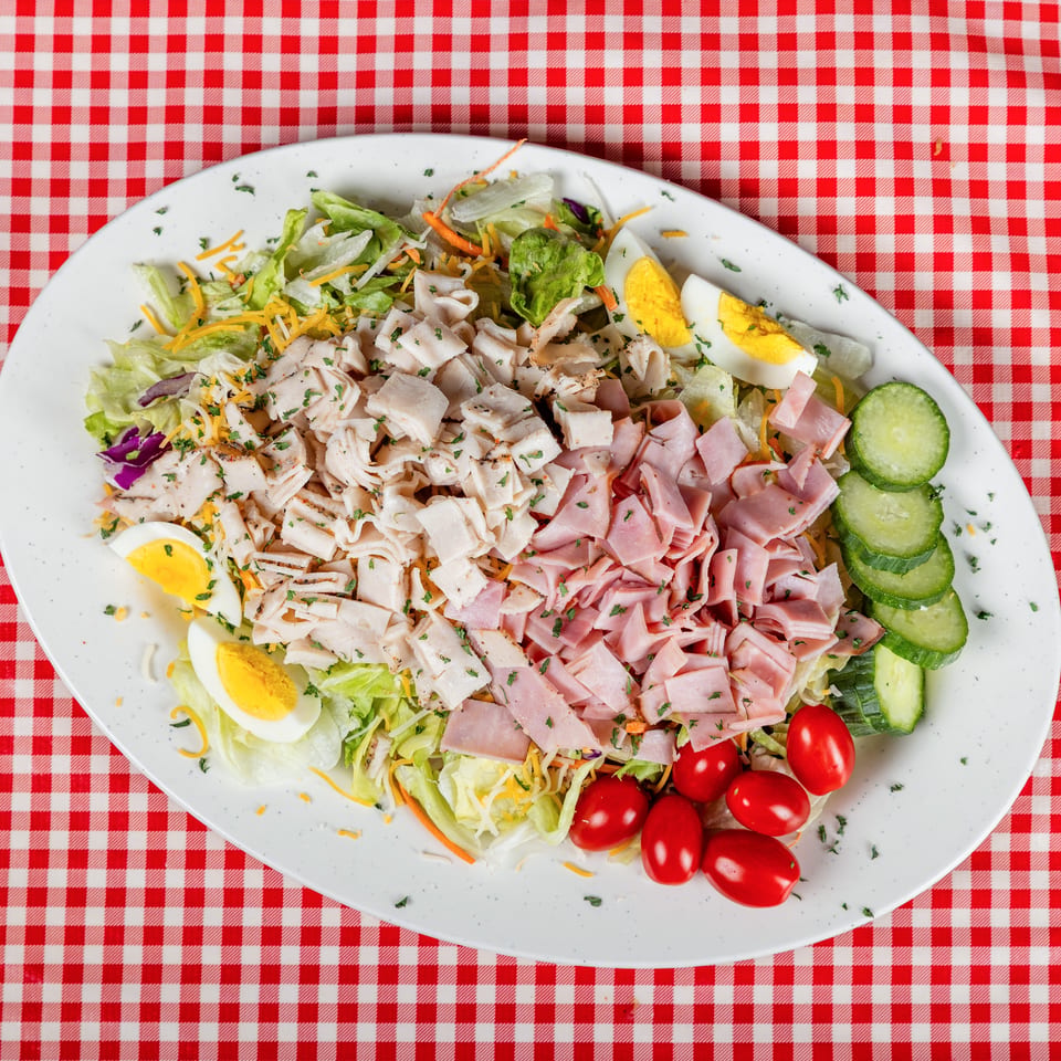 Chef salad with turkey, ham, lettuce, cheddar, hard-boiled eggs, cucumbers, and tomatoes on a red-and-white checkered tablecloth.