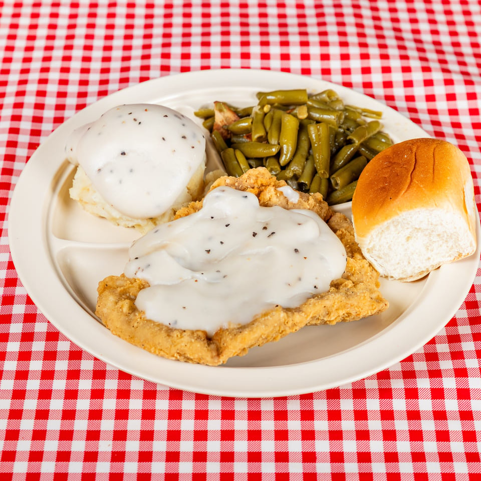 Southern-style fried chicken smothered in peppered gravy with mashed potatoes, green beans, and a dinner roll on a checkered tablecloth.