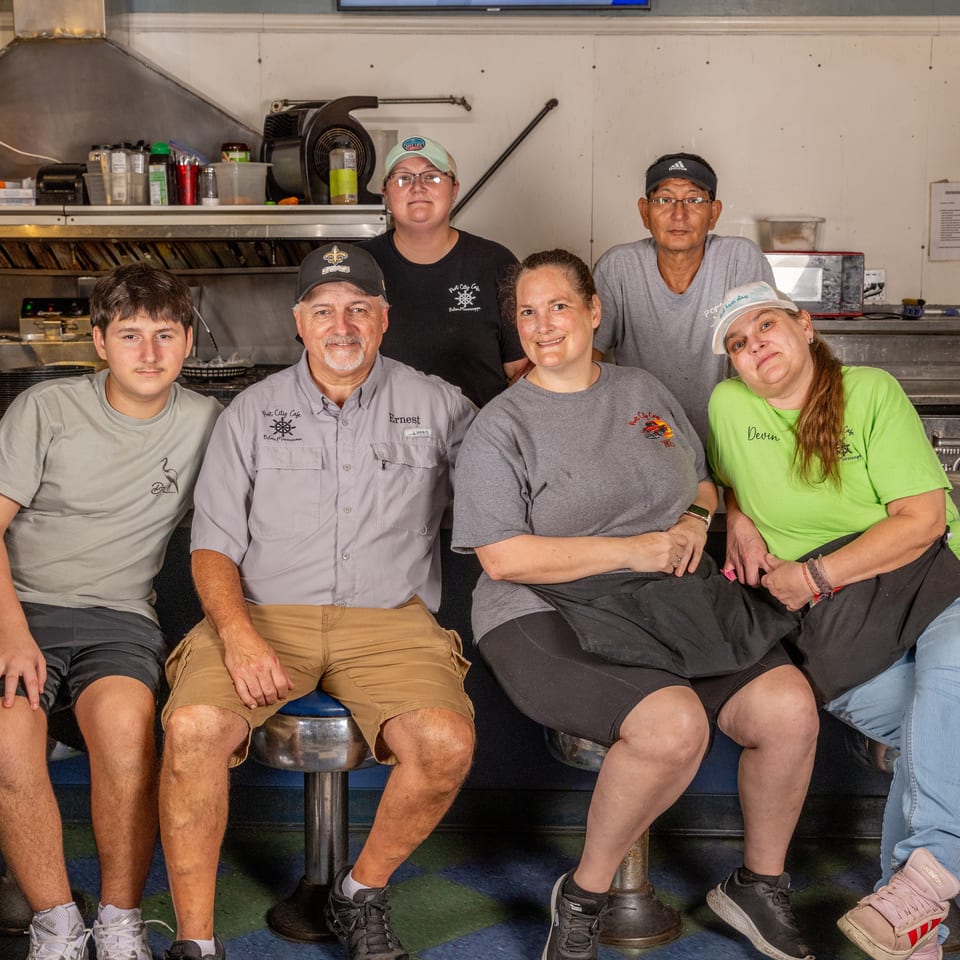 Six people pose together inside a restaurant kitchen, seated and standing near the counter and cooking equipment.