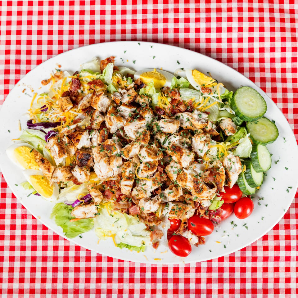 Grilled chicken and bacon salad with lettuce, cheddar cheese, eggs, cucumbers, tomatoes, and herbs on a red-checkered tablecloth.