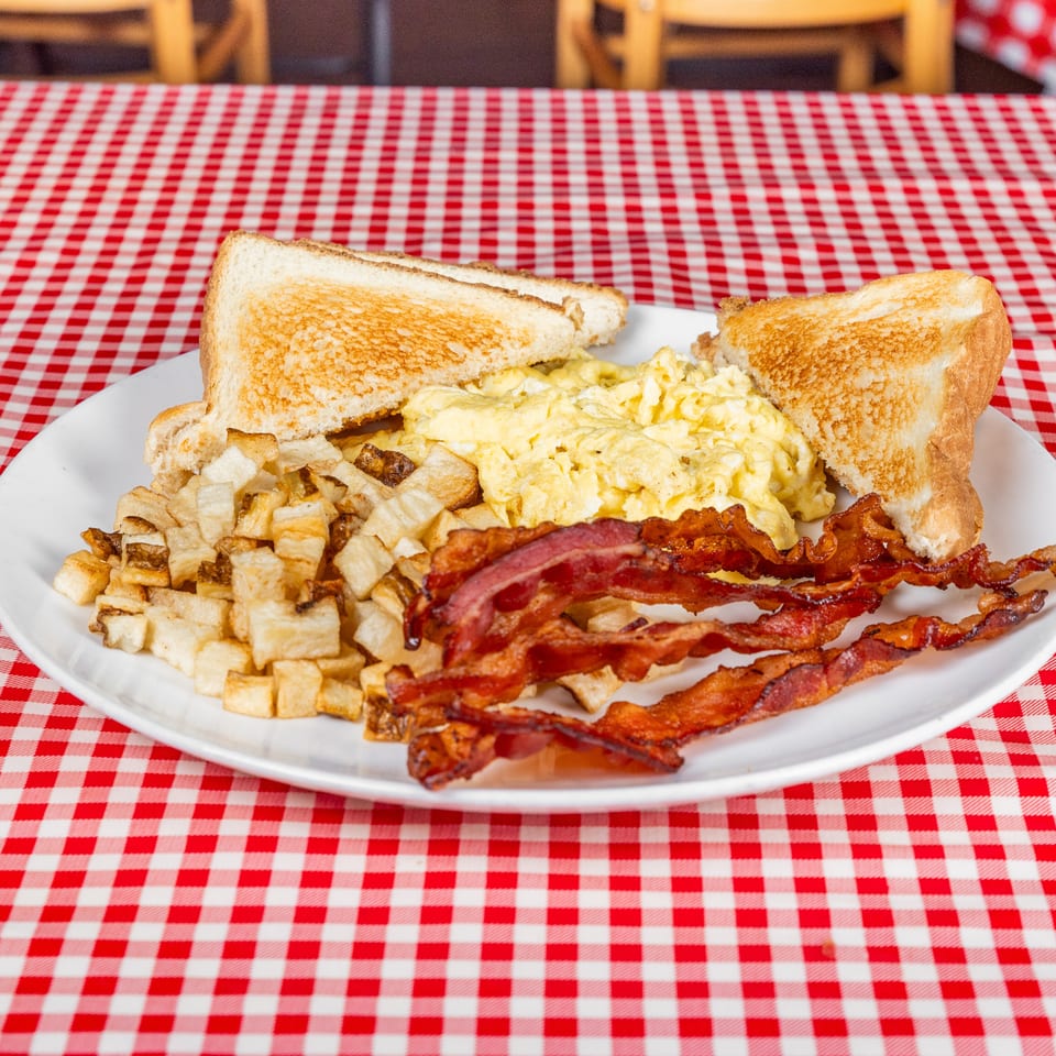 Plate of scrambled eggs, toast, home fries, and crispy bacon on a red-and-white checkered diner tablecloth.