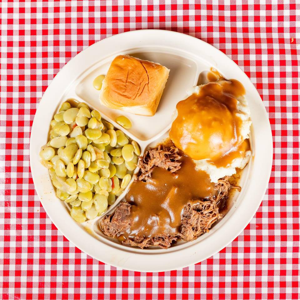 Plate of pulled pork with gravy, mashed potatoes, butter beans, and a dinner roll on a red-and-white checkered tablecloth.