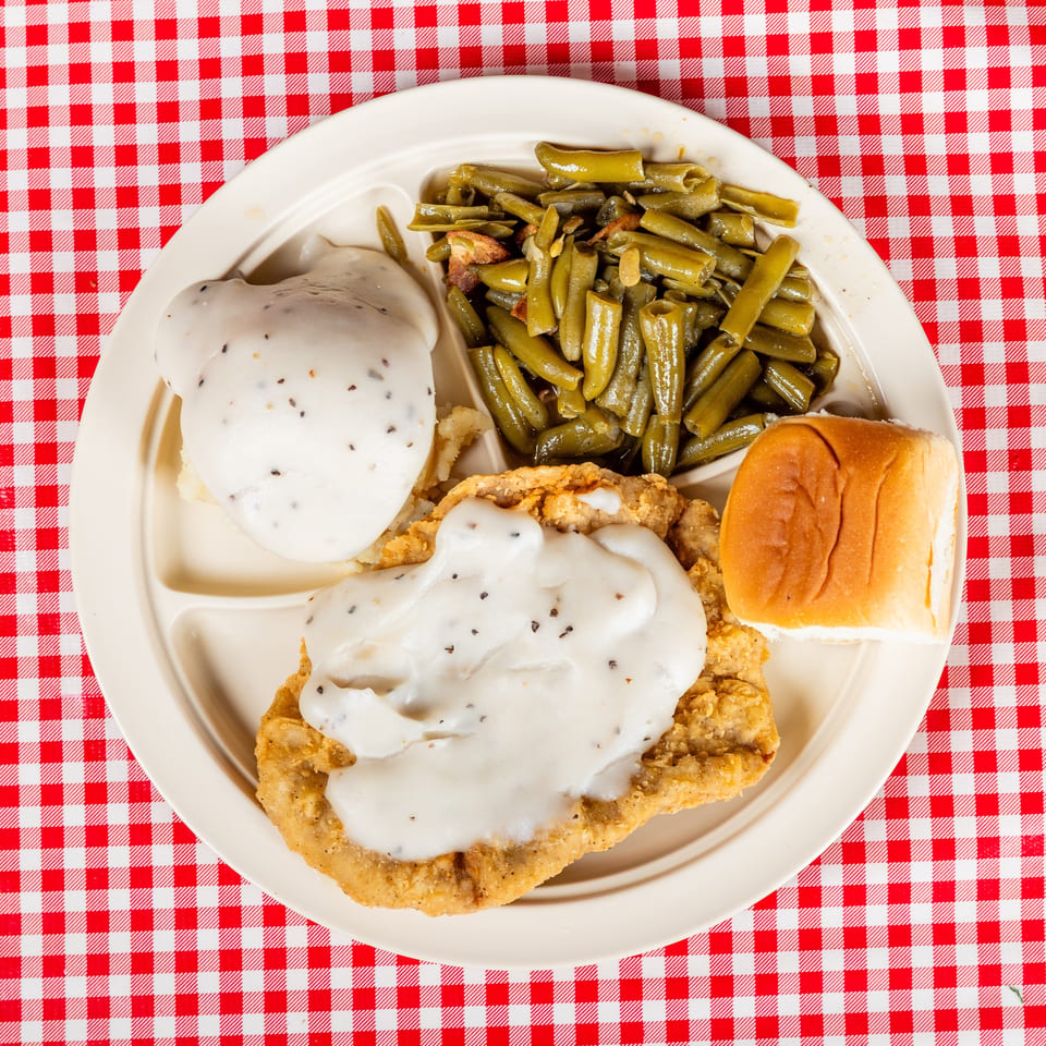 Southern-style plate with fried chicken covered in white gravy, mashed potatoes, green beans, and a dinner roll on red-checkered tablecloth.