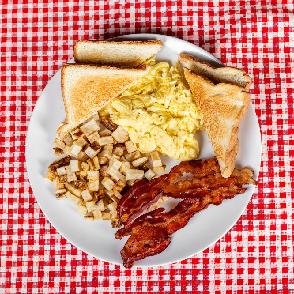 Plate of scrambled eggs, toast, breakfast potatoes, and bacon on a red-and-white checkered tablecloth.
