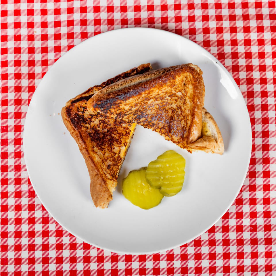 Grilled triangular sandwich with a bite taken out, served with pickle slices on a white plate atop a red-and-white checkered tablecloth.