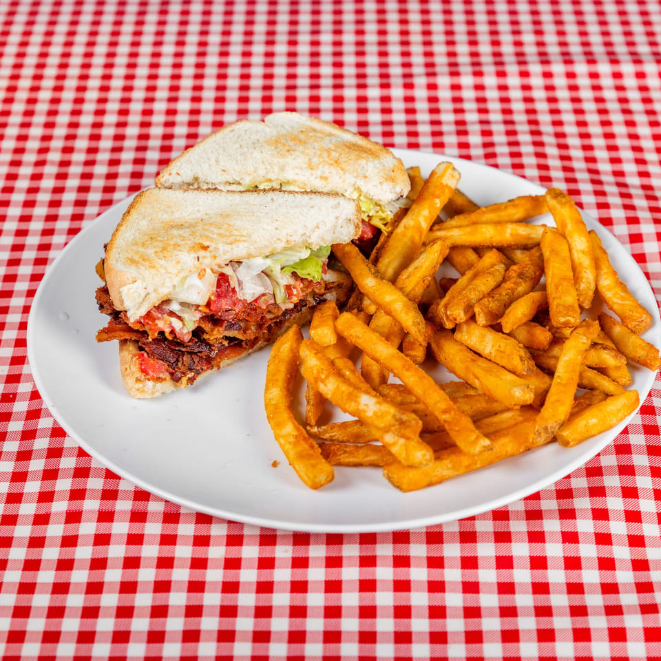 Toasted sandwich with lettuce, tomato, and bacon served alongside golden fries on a white plate with red checkered tablecloth.