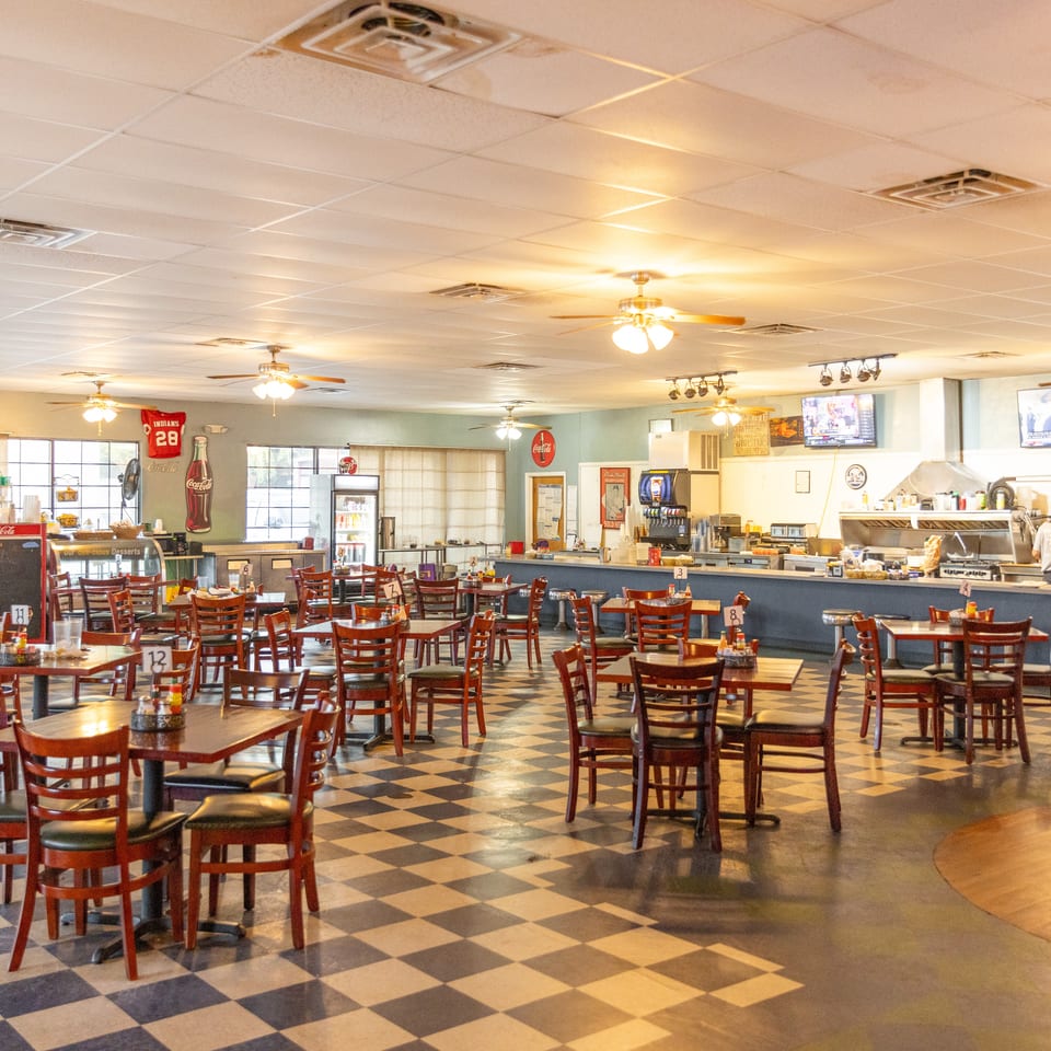 Spacious diner interior with checkerboard floors, wooden tables, counter seating, televisions, and a visible open kitchen.