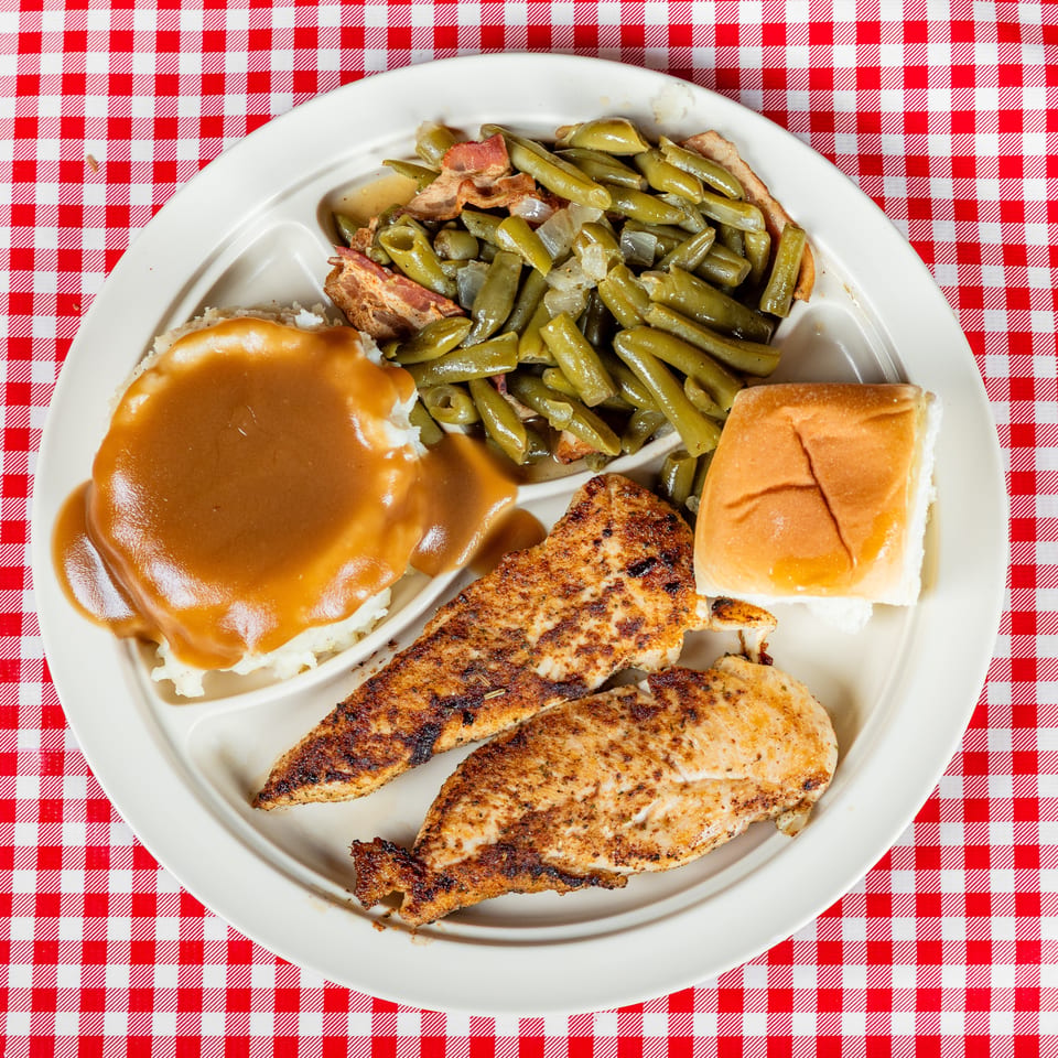 Southern-style plate with grilled chicken, mashed potatoes and gravy, green beans with bacon, and a dinner roll on a red checkered tablecloth.