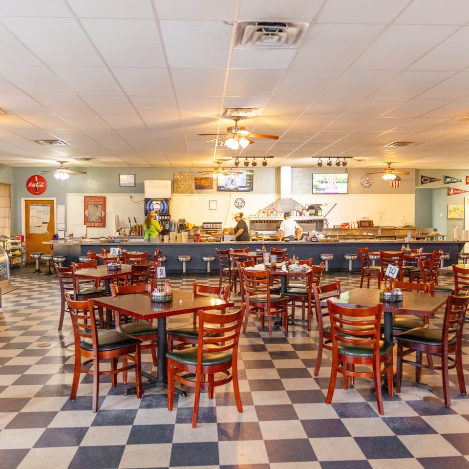 Spacious casual restaurant dining room with wooden tables, red chairs, checkered floor, sports memorabilia, televisions, and an open service counter.