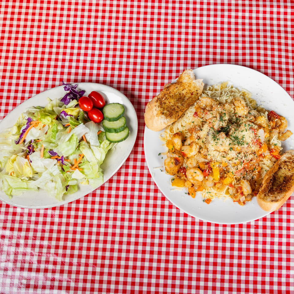 A plate of shrimp pasta with seasoned bread accompanies a fresh garden salad on a red-and-white checkered tablecloth.
