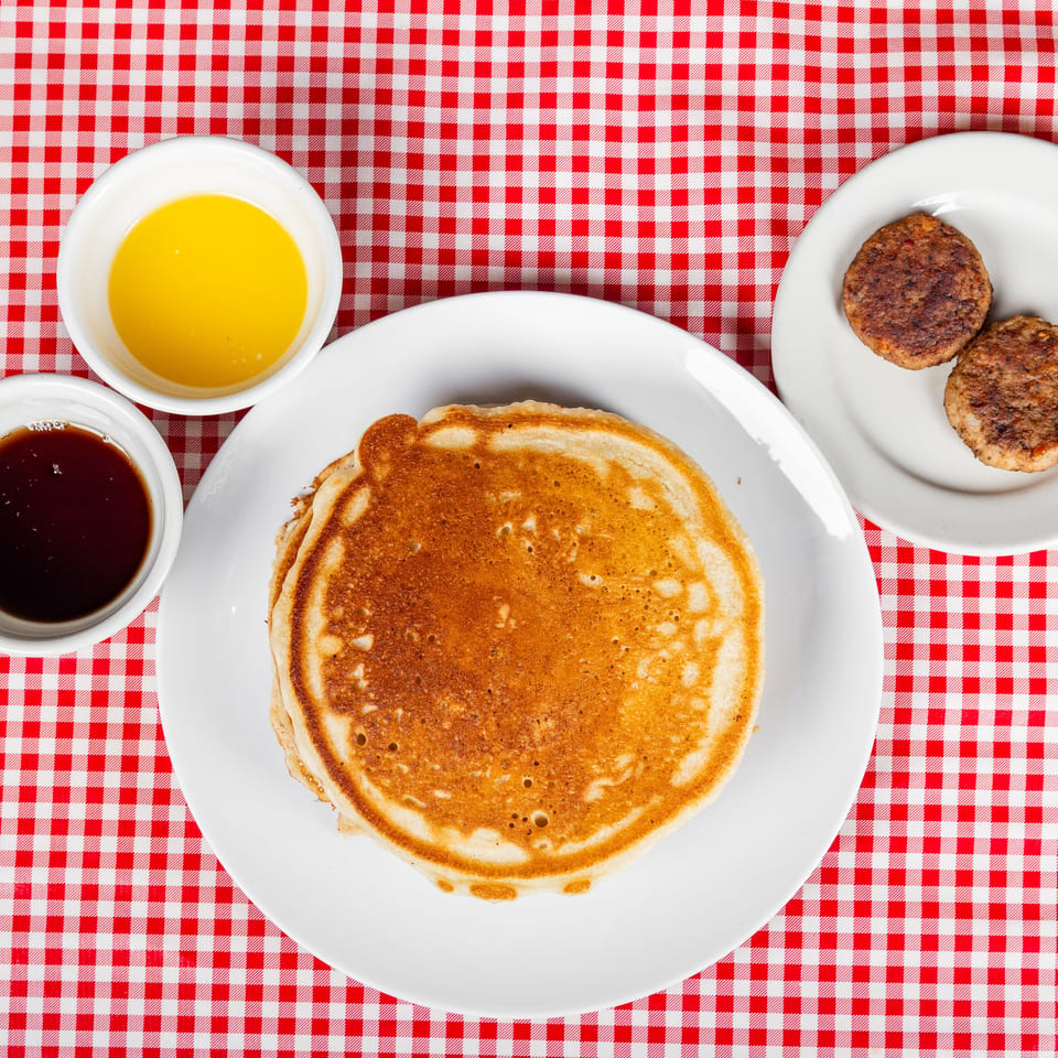 Stack of pancakes with syrup, butter, and two sausage patties on white plates over a red-and-white checkered tablecloth.